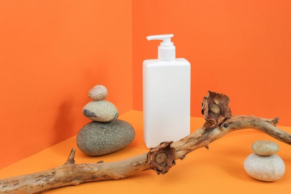 One white blank cosmetic bottle with dispenser, rocks, wooden stick with dried flowers in corner space on orange background. Mockup Front view Copy space.